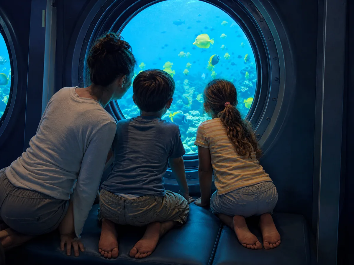 Bambini e un genitore che guardano da un oblò rotondo del sottomarino l'acqua blu e i pesci gialli